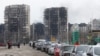 Uma fila de carros com pano de fundo prédios residenciais destruídos pelo conflito Ucrânia-Rússia. Residentes tentam sair da cidade portuária de Mariupol, Ucrânia, 17 Março, 2022.